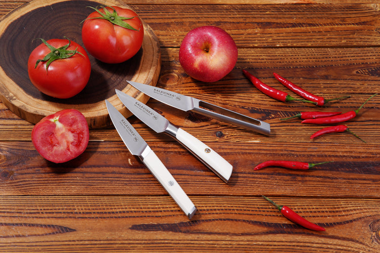 Three kitchen knives with white handles are arranged on a wooden surface, along with two tomatoes on a wooden board, one sliced tomato, an apple, and several red chili peppers.