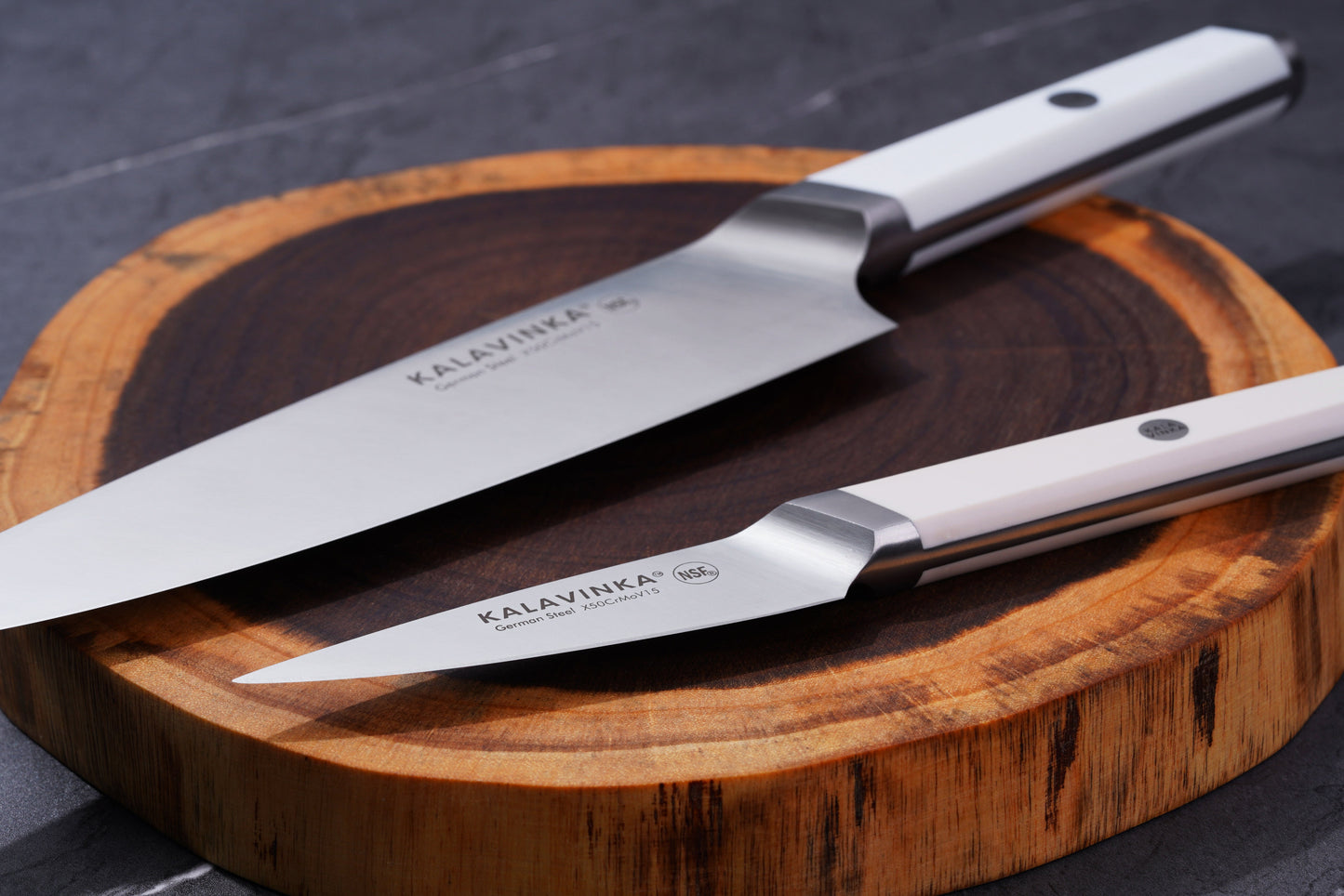 Two stainless steel knives with white handles rest elegantly on a wooden cutting board. The knives bear the brand name "KALAVINKA" on their blades, one being a larger chef's knife and the other a smaller paring knife. The background is a dark grey surface.