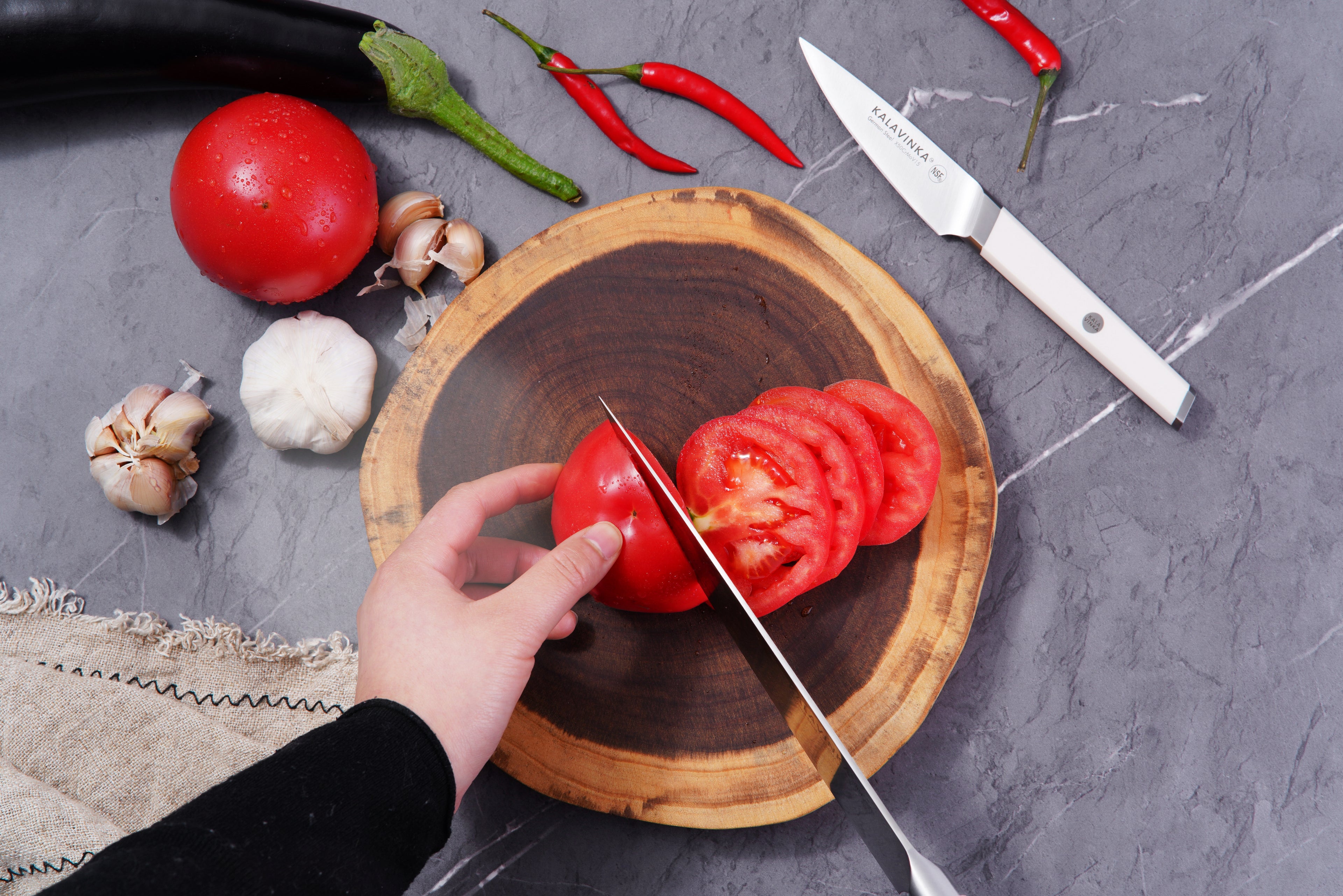 A hand slicing a tomato on a wooden cutting board with a knife. Surrounding the plate are whole tomatoes, garlic, red chili peppers, and an eggplant. Another paring knife rests on the gray marble countertop.
