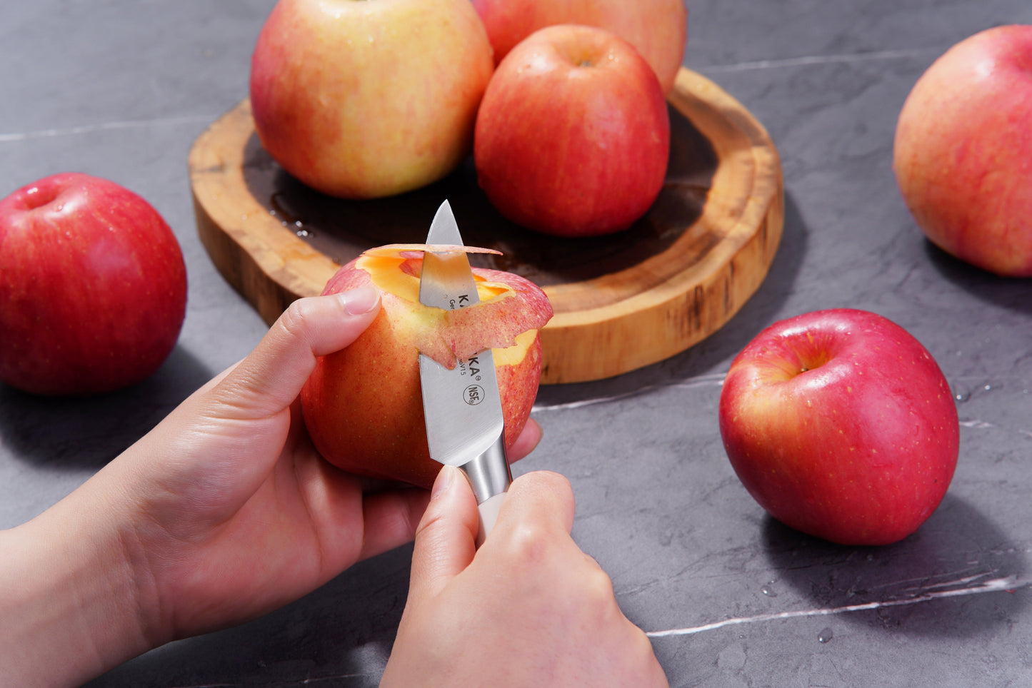 A person uses a paring knife to peel a red apple, with more apples on a wooden plate and table in the background.