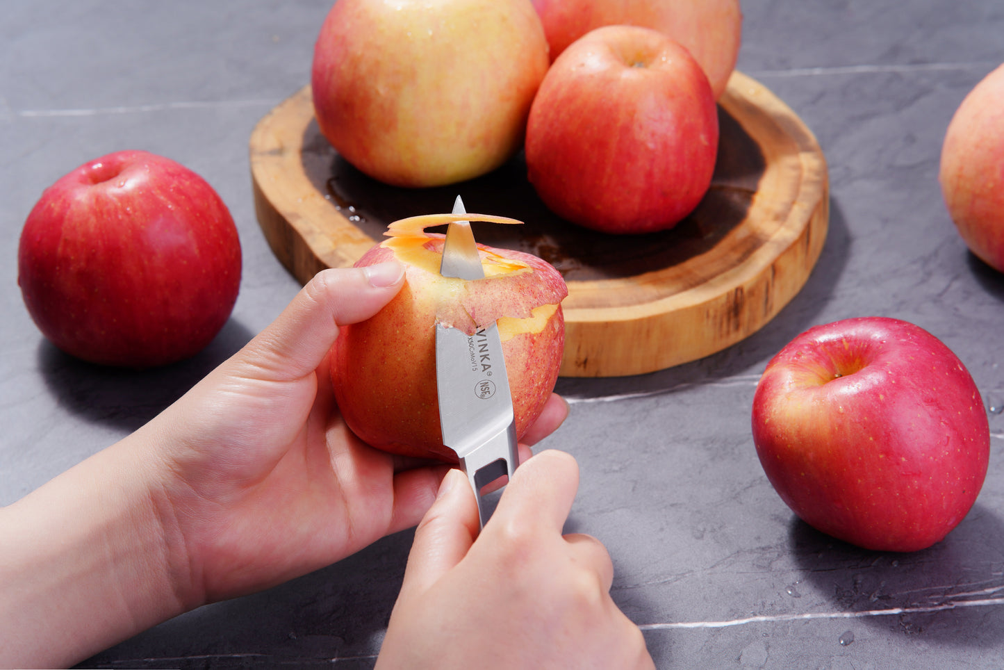 Using the KALAVINKA VENDANA Series 2-Piece Essentials Knife Set, a person peels a red apple with the precision of the 3.5-Inch Paring Knife over a gray countertop. Several other apples are scattered around, with some resting on a wooden cutting board in the background. The focus is on the apple being expertly peeled using high-carbon steel craftsmanship.