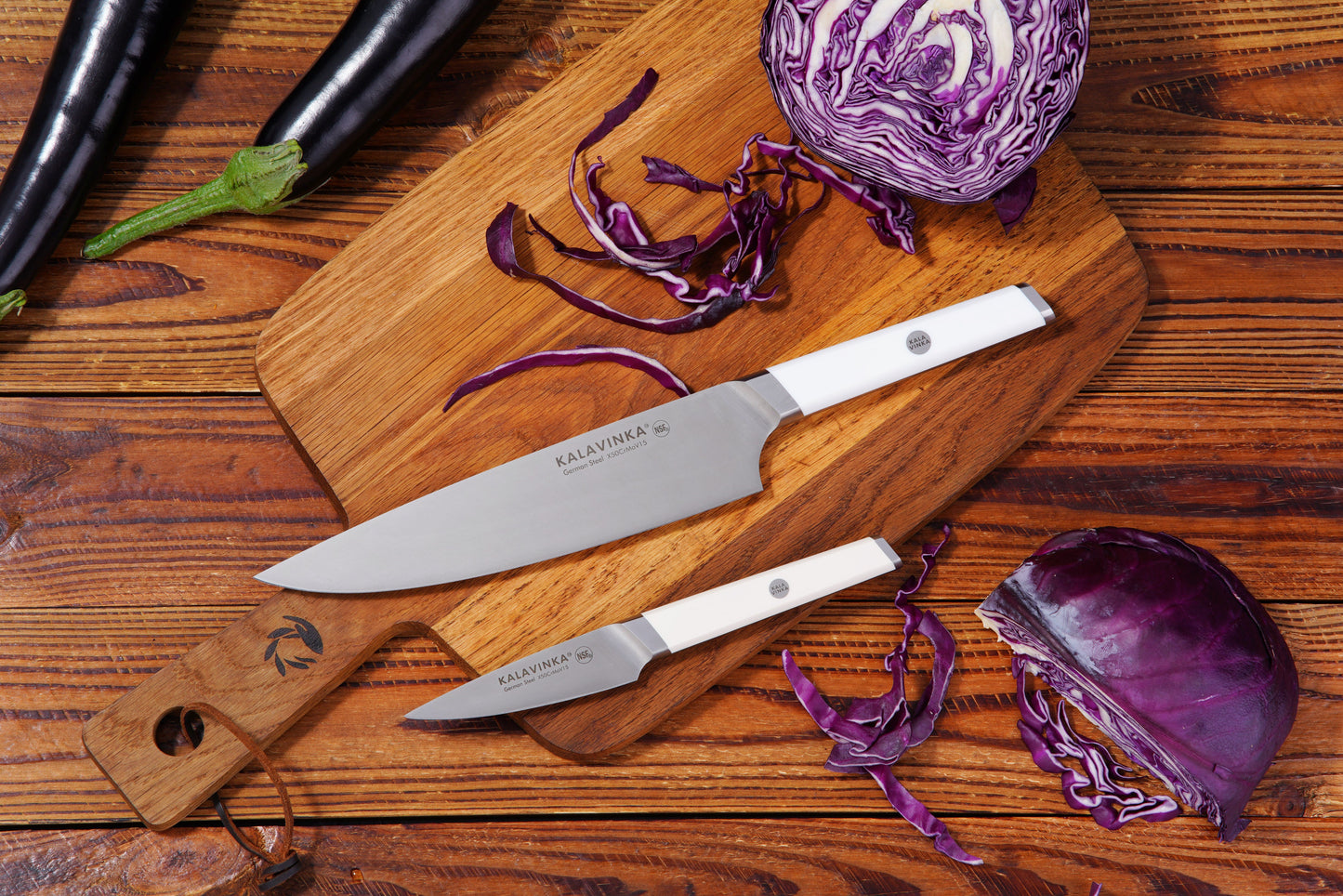 Two knives on a wooden cutting board with chopped red cabbage and whole eggplants.