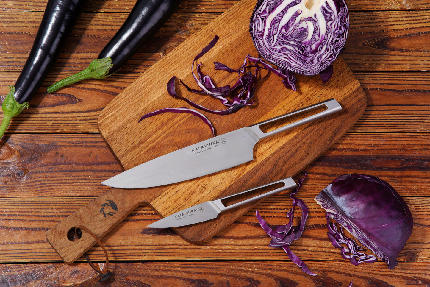 A wooden cutting board with the VENDANA Series 2-Piece Essentials Knife Set from KALAVINKA, featuring an 8-inch chef's knife and a 3.5-inch paring knife, lies on a wooden surface. Purple cabbage is sliced on the board, with additional pieces scattered around. Two whole eggplants sit nearby, adding a splash of green to the otherwise vibrant purple composition.