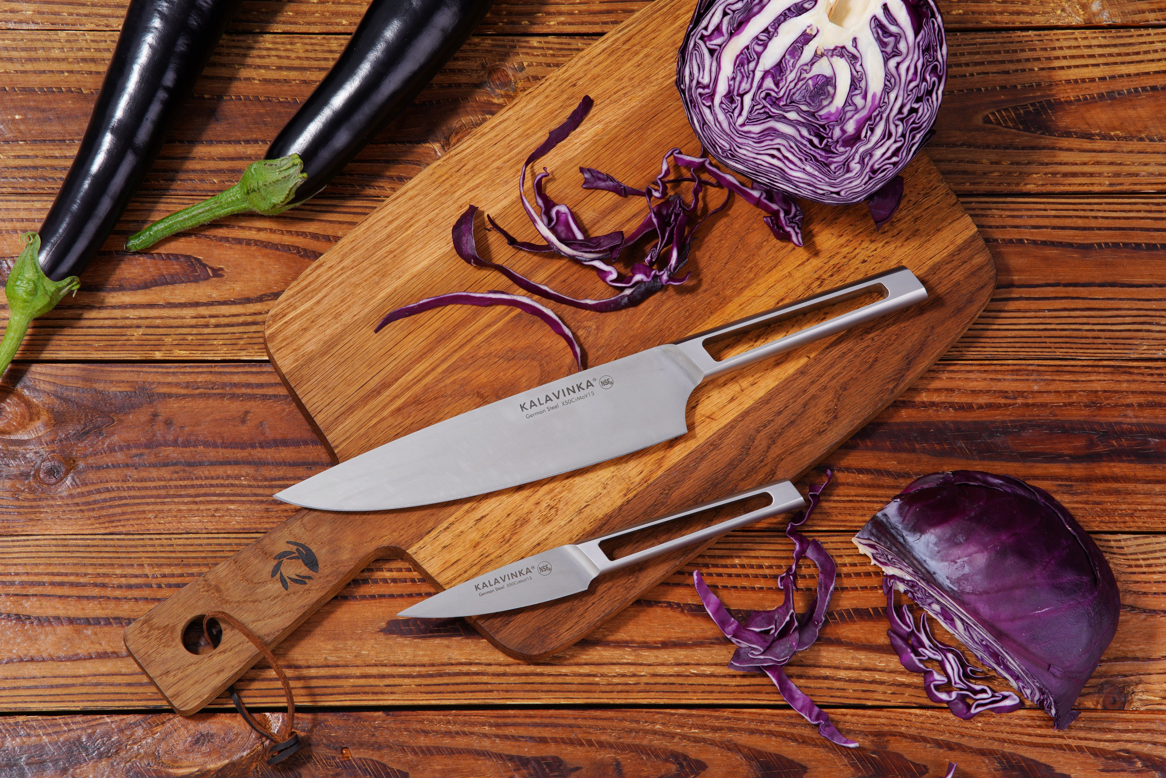 A wooden cutting board with the VENDANA Series 2-Piece Essentials Knife Set from KALAVINKA, featuring an 8-inch chef's knife and a 3.5-inch paring knife, lies on a wooden surface. Purple cabbage is sliced on the board, with additional pieces scattered around. Two whole eggplants sit nearby, adding a splash of green to the otherwise vibrant purple composition.