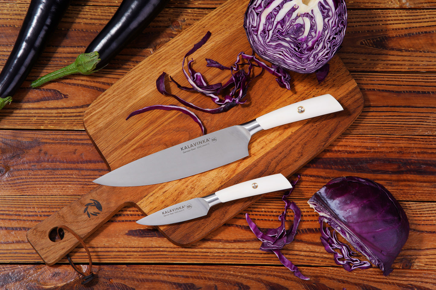 Two knives on a wooden cutting board with chopped red cabbage and whole eggplants.