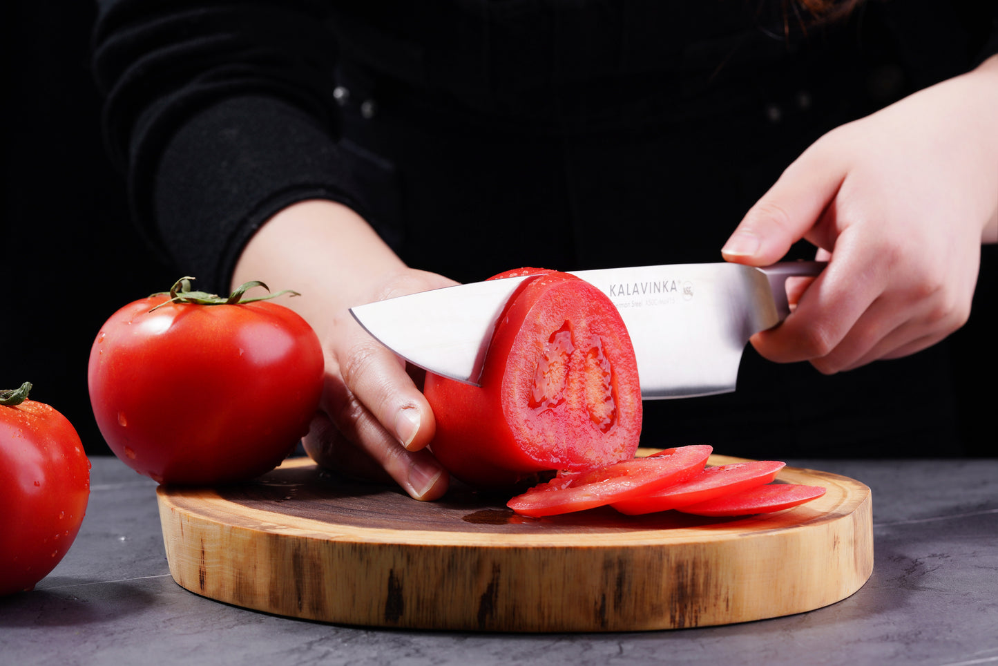 Close-up of a person slicing a ripe tomato with the 8-Inch Chef's Knife from the VENDANA Series 2-Piece Essentials Knife Set by KALAVINKA on a wooden cutting board. Two whole tomatoes are on the board, and several tomato slices are neatly arranged. The person wears a dark top, and the background is black.