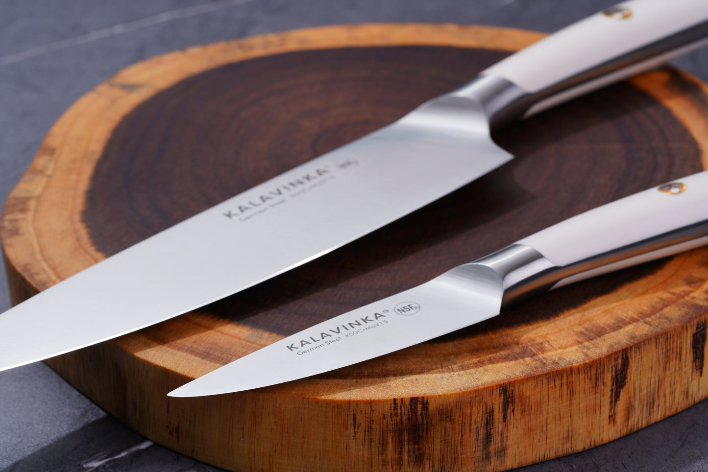 Two stainless steel knives with white handles rest elegantly on a wooden cutting board. The knives bear the brand name "KALAVINKA" on their blades, one being a larger chef's knife and the other a smaller paring knife. The background is a dark grey surface.