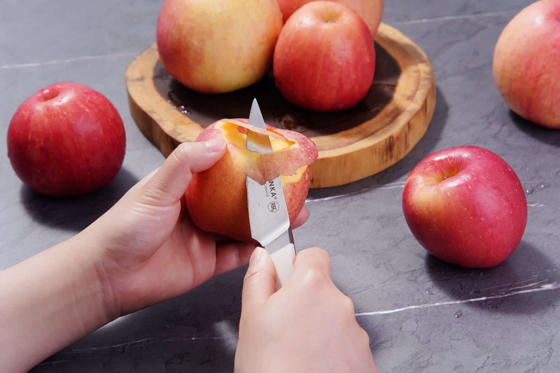 A person uses a paring knife to peel a red apple, with more apples on a wooden plate and table in the background.
