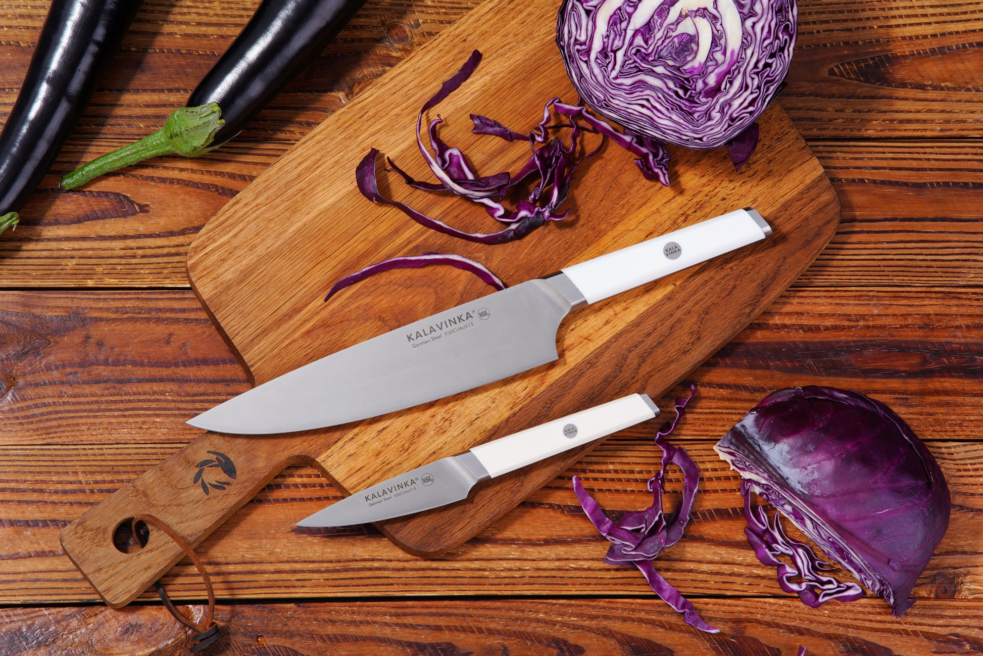 Two knives on a wooden cutting board with chopped red cabbage and whole eggplants.