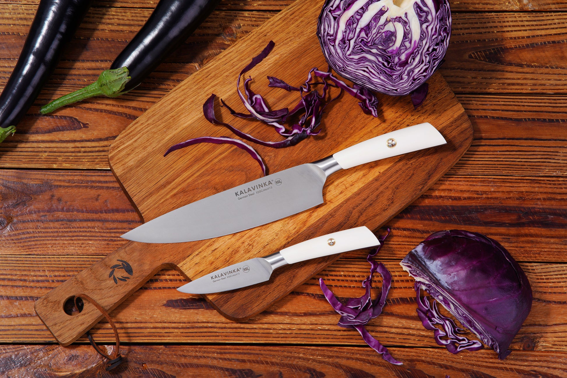 Two knives on a wooden cutting board with chopped red cabbage and whole eggplants.